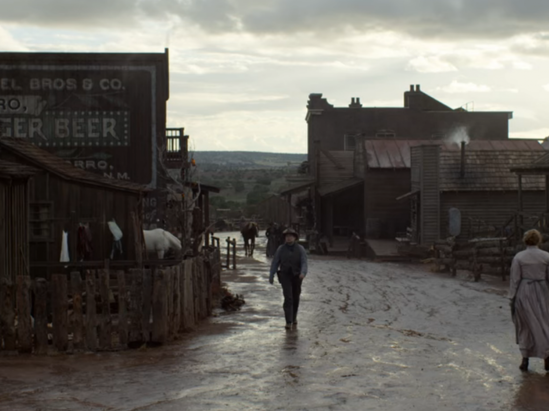 Szenenbild aus der Serie. Man sieht den Eingang eines Western-Dorfes. Der Weg ist schlammig, der Himmel düster und zugezogen.