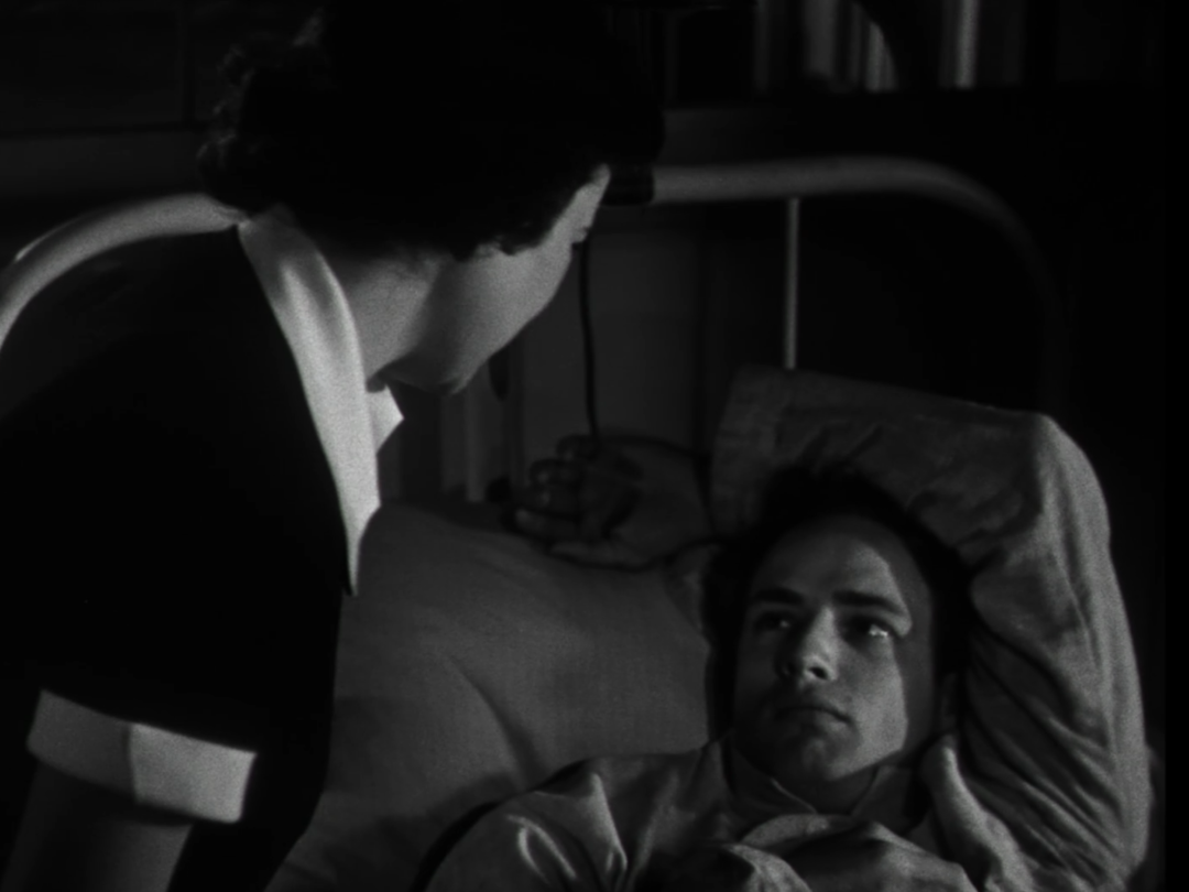 Das Bild zeigt die Figuren Ellen und Ken aus dem Film "Die Männer" von 1950. Ellen sitzt an Kens Bett, die beiden sehen sich an. Das Bild ist schwarz-weiß und der Raum sehr dunkel.