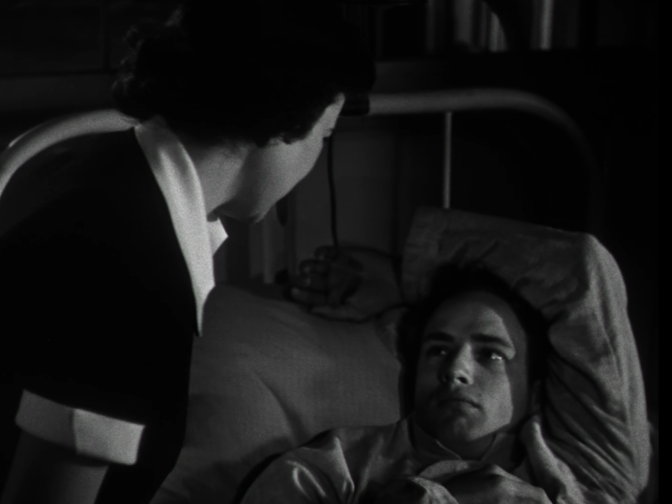 Das Bild zeigt die Figuren Ellen und Ken aus dem Film "Die Männer" von 1950. Ellen sitzt an Kens Bett, die beiden sehen sich an. Das Bild ist schwarz-weiß und der Raum sehr dunkel.