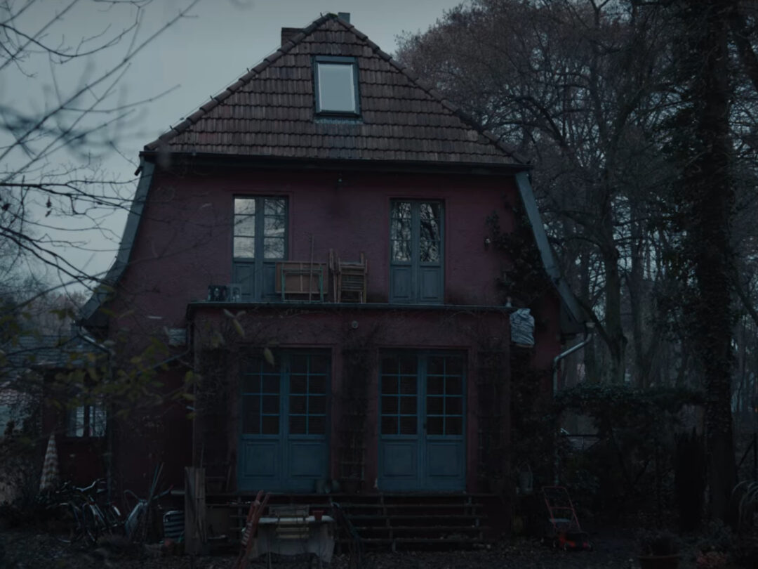 Ein Screenshot aus der Serie "Dark". Man sieht ein dunkelrot gestrichenes Haus, daneben schwarze Silhouetten von Bäumen. Es regnet und der Himmel ist sehr dunkel.