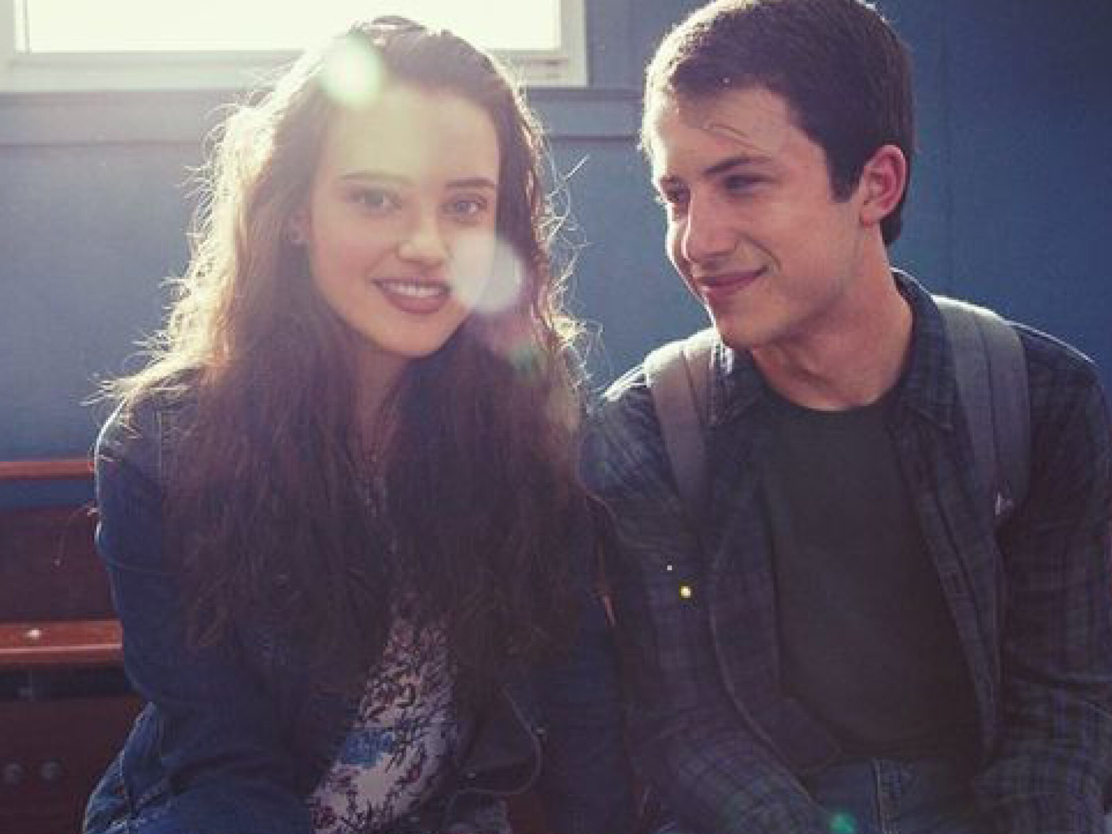 Auf dem Bild sieht man die beiden Hauptfiguren der Serie Hannah und Clay. Sie sitzen nebeneinander und lächeln sich an. Das Licht fällt durch ein Fenster hinter Hannah und lässt ihre Haare strahlen wie einen Heiligenschein.